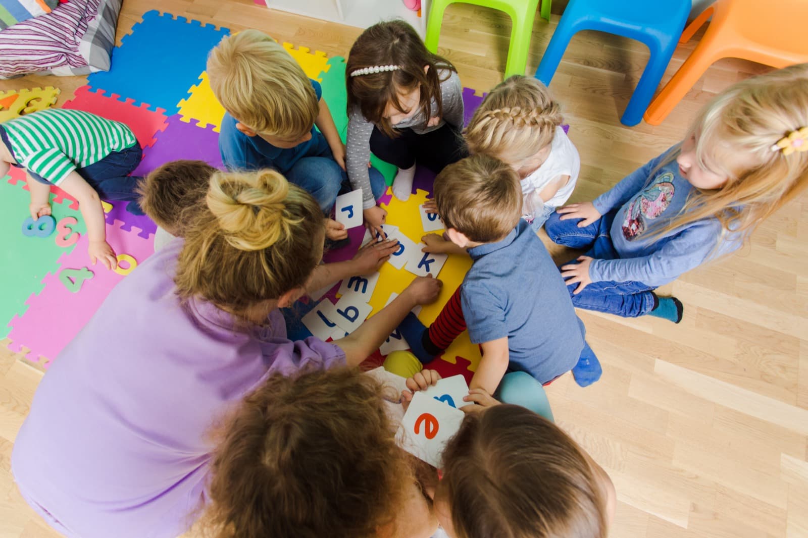 Kinder und Erzieherin spielen gemeinsam auf bunter Matte mit Buchstabenkarten